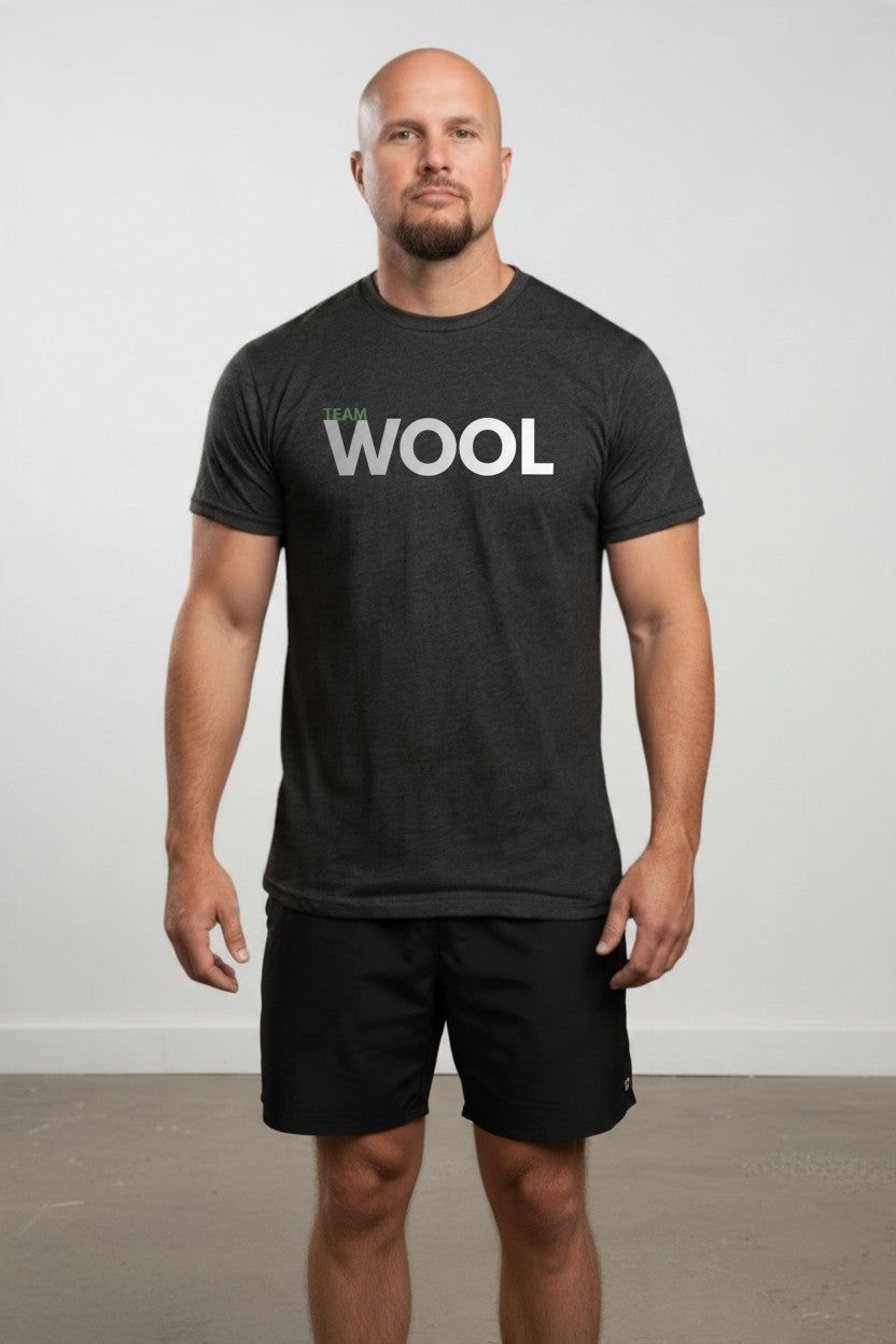A man wearing a dark charcoal gray t-shirt with 'team WOOL' printed in white across the chest, paired with black athletic shorts, standing against a neutral light gray studio background.