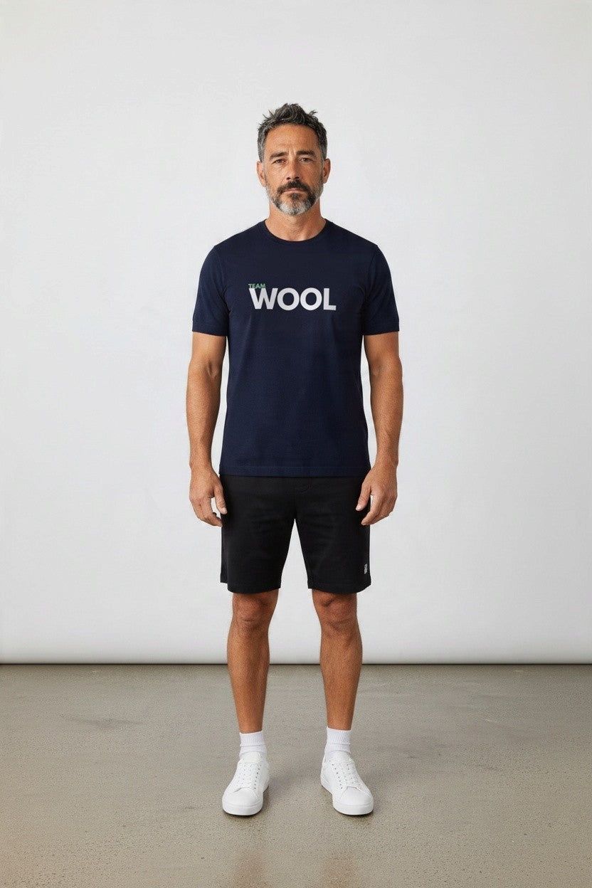 A man stands facing forward in a studio wearing a navy blue t-shirt with 'WOOL' printed in white across the chest, black shorts, white socks, and white sneakers against a light gray wall and concrete floor.