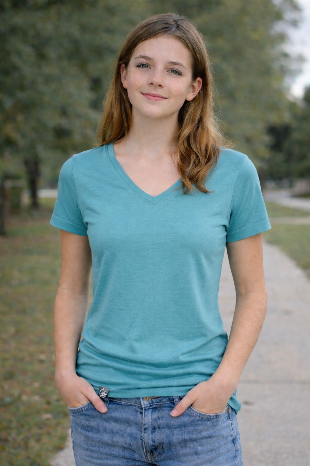 Woman wearing a teal t-shirt and blue jeans standing outdoors with trees in the background